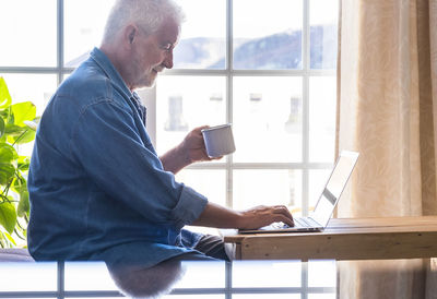 Side view of man using laptop while drinking coffee at home