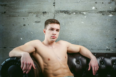 Shirtless muscular man sitting on armchair by wall