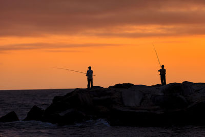 Silhouette man fishing on rock by sea against sky during sunset