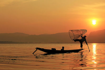 Silhouette fisherman holding cone shaped fishing net while standing on boat in lake during sunset