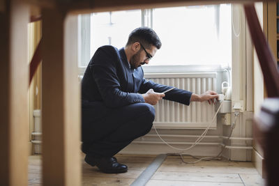 Lawyer crouching while charging phone by radiator with furniture in foreground