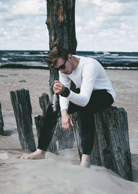 Full length of young man falling sand while sitting on wooden post at beach