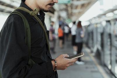Midsection of man using smart phone while standing at railroad platform