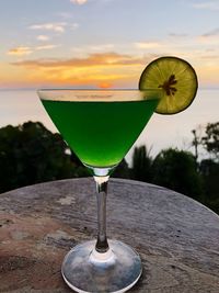 Green drink in glass on table against sea during sunset