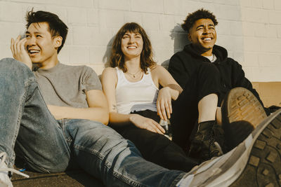 Smiling male and female friends sitting on footpath against wall during sunny day