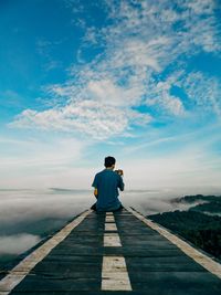 Rear view of man photographing cloudscape over mountain while sitting on jetty against sky