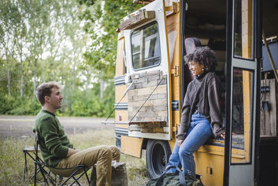 Smiling woman leaning on caravan doorway while talking with male friend sitting during camping in forest