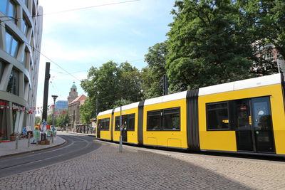 Yellow train by trees against sky in city