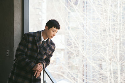 Young man looking away while standing on window