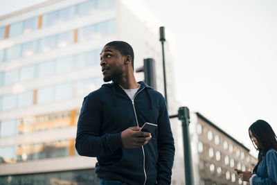 Low angle view of young man standing with mobile phone while looking away against building in city