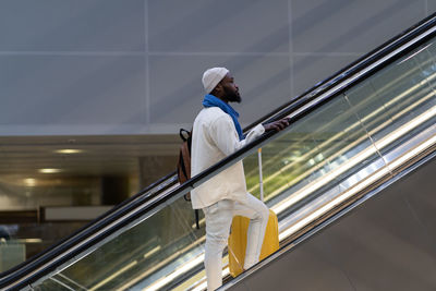 African american passenger man with suitcase stands on escalator, holds handrail in airport terminal