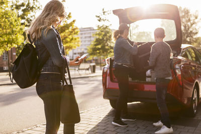 Side view of teenager using phone while mother and son keeping suitcase in car trunk