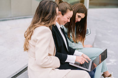 Side view of company of smiling businesswomen in smart casual clothes sitting in city and using laptop while working on startup project together